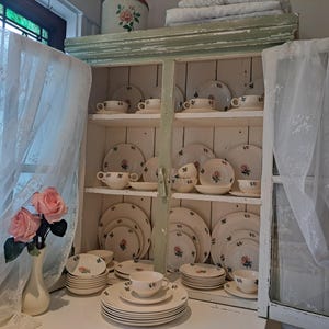 May include: A white cabinet displays a collection of vintage dishware. The plates, bowls, and cups feature a delicate pink rose design. The cabinet is painted in a distressed green, and the scene is complemented by sheer curtains and a vase of pink roses.