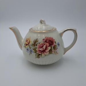 May include: Cream-colored ceramic teapot with a lid, handle, and spout. The teapot is decorated with a floral design featuring pink, yellow, and blue roses. Gold trim accents the edges of the lid, spout, and handle.