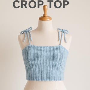 May include: A light blue crochet crop top with tie straps. The top is displayed on a mannequin and features the text "CROP TOP" at the top of the image. The crochet stitch creates a textured appearance.