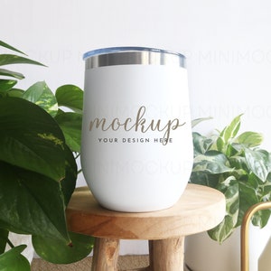 May include: White stainless steel tumbler with a silver lid and a gold inscription that reads "mockup your design here". The tumbler is sitting on a wooden stool.