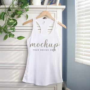 May include: White tank top with a racerback style hanging on a wooden hanger on a white dresser. The tank top has the word "mockup" written on it in brown text.