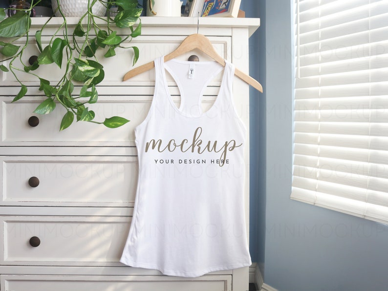 May include: White tank top with a racerback style hanging on a wooden hanger on a white dresser. The tank top has the word "mockup" written on it in brown text.