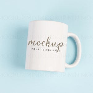 May include: White ceramic mug with a handle on a light blue background. The mug has the word "mockup" written on it in brown cursive font with the text "Your design here" underneath.