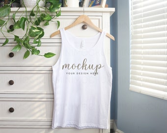 May include: White tank top mockup hanging on a wooden hanger on a white dresser with a green plant in the background. The tank top has the text "mockup your design here" printed on the front.