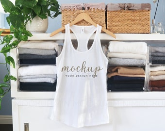 May include: White tank top mockup with the text "mockup your design here" on a white shelf with folded clothing in clear containers.