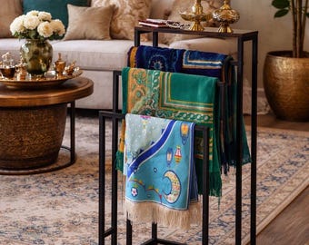 Op de afbeelding: Een zwart metalen rek met verschillende gebedskleden met ingewikkelde patronen en franjes. De kleden zijn in tinten blauw, groen en turquoise. Het rek staat in een woonkamer met een bank, salontafel en decoratieve items.