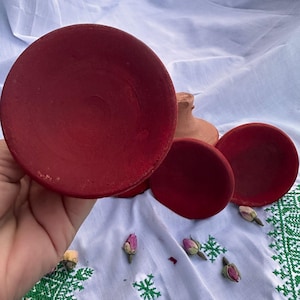 May include: Three circular terracotta dishes with a matte red finish on the interior and a natural clay exterior. The dishes are shallow with a smooth, rounded rim. One dish is held in the foreground, while two others sit behind it.