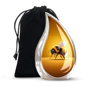 May include: A silver-colored, teardrop-shaped urn with a detailed bee illustration on a yellow background. The urn is accompanied by a black velvet drawstring bag. The urn has a decorative etched pattern around the edges.