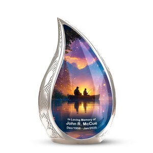 May include: A teardrop-shaped silver cremation urn featuring a colourful printed scene of two people fishing in a boat on a lake at sunset. The text reads In Loving Memory of John R. McCue Dec/1956 - Jan/2025 with a textured silver border.
