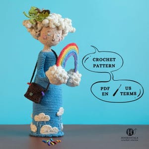 Può includere: Bambola all'uncinetto fatta a mano con un vestito blu, che tiene un arcobaleno e dettagli di nuvole bianche. La bambola ha una borsa marrone e una rana verde sulla testa. Il testo sull'immagine dice "CROCHET PATTERN" e "PDF EN US TERMS".