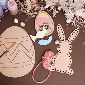 May include: A craft kit for making Easter decorations. It includes a wooden egg with a chevron pattern, a wooden bunny with holes for stringing yarn, a wooden egg with the words "Say our Easter wishes", and a small paintbrush. The kit also includes paint, yarn, and small plastic eggs.
