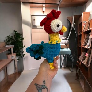 May include: A crocheted rooster toy with a red comb, yellow beak, blue body, and green tail feathers. The toy is being held in a person's hand.