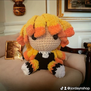 May include: A crocheted plush toy of a character with yellow and orange hair, wearing a black and white outfit. The toy is sitting on a beige sofa.