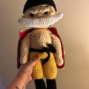 May include: A handmade crochet doll featuring a character with a black bandana, a large white mustache, and a tan complexion. The figure wears yellow pants with a black sash, black boots, and a red cape with gold trim draped over the shoulders.
