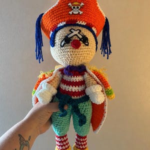 May include: A crocheted doll featuring a beige head with blue hair, a red nose, and a large orange pirate hat adorned with a skull and crossbones emblem. The doll wears a red and white striped shirt, teal pants, white gloves, and a colorful cape.