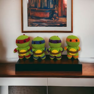 May include: Four crocheted Teenage Mutant Ninja Turtles, each with a different colored mask: red, purple, blue, and orange. The turtles are standing on a black shelf.