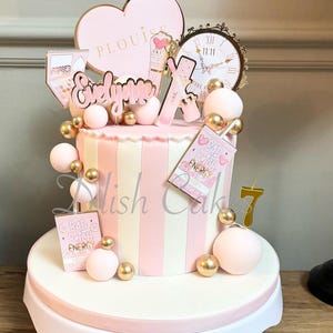 May include: A striped cake in pink and white, adorned with gold and pink spheres, and decorative toppers. The toppers include a heart, a clock, and the name "Evelynne". The cake is presented on a white cake stand.