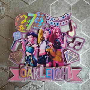 Puede incluir: Un adorno para pastel rosa y plateado con tres figuras de dibujos animados sosteniendo micr&oacute;fonos. El adorno incluye el texto "OAKLEIGH", el n&uacute;mero "7" y las palabras "KPOP DEMON HUNTERS". Tambi&eacute;n se incluye una bola de discoteca y notas musicales.
