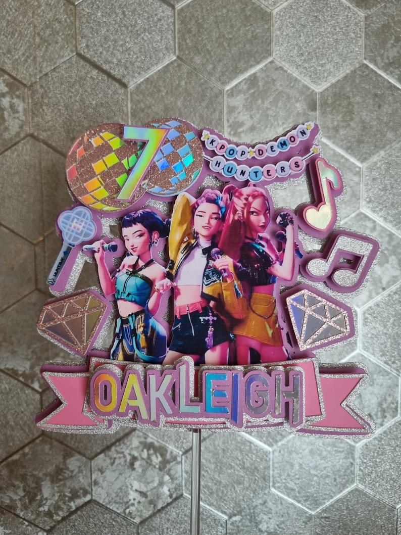 Puede incluir: Un adorno para pastel rosa y plateado con tres figuras de dibujos animados sosteniendo micr&oacute;fonos. El adorno incluye el texto "OAKLEIGH", el n&uacute;mero "7" y las palabras "KPOP DEMON HUNTERS". Tambi&eacute;n se incluye una bola de discoteca y notas musicales.