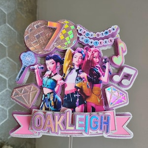 Puede incluir: Un adorno para tarta rosa y plateado con tres figuras de dibujos animados sosteniendo micr&oacute;fonos. El adorno incluye el texto "OAKLEIGH", el n&uacute;mero "7" y las palabras "KPOP DEMON HUNTERS". Tambi&eacute;n se incluyen notas musicales y diamantes decorativos.