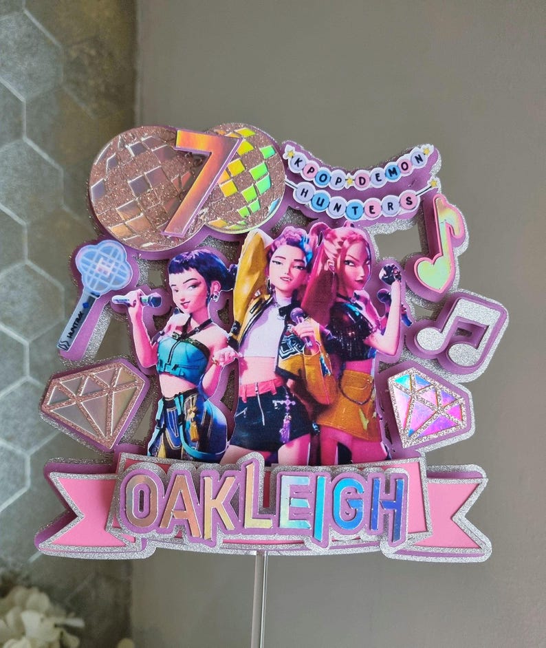 Puede incluir: Un adorno para tarta rosa y plateado con tres figuras de dibujos animados sosteniendo micr&oacute;fonos. El adorno incluye el texto "OAKLEIGH", el n&uacute;mero "7" y las palabras "KPOP DEMON HUNTERS". Tambi&eacute;n se incluyen notas musicales y diamantes decorativos.