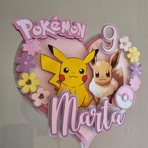 May include: A pink heart-shaped cake topper with a pink and white Pok&eacute; Ball, a yellow Pikachu, a brown Eevee, and the text "Pok&eacute;mon" and "Marta 9".