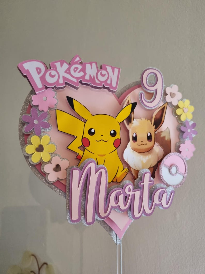May include: A pink heart-shaped cake topper with a pink and white Pok&eacute; Ball, a yellow Pikachu, a brown Eevee, and the text "Pok&eacute;mon" and "Marta 9".