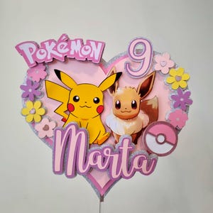 May include: A pink heart-shaped cake topper with a pink and silver glitter finish. The topper features the Pokemon characters Pikachu and Eevee, the name "Marta", and the number "9".