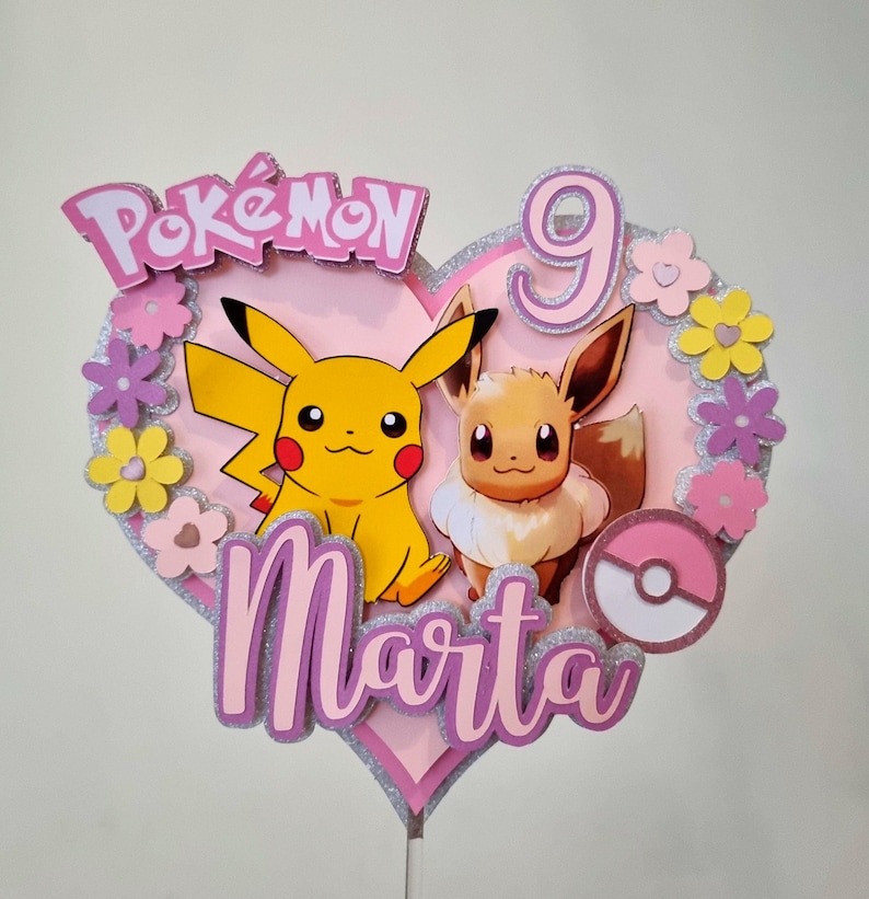 May include: A pink heart-shaped cake topper with a pink and silver glitter finish. The topper features the Pokemon characters Pikachu and Eevee, the name "Marta", and the number "9".