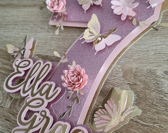 May include: A pink and gold glitter cake topper with the name "Ella Grace" and a number 7 decorated with butterflies and flowers.