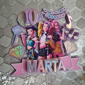 Puede incluir: Un adorno para tarta con tres figuras femeninas estilizadas sosteniendo micr&oacute;fonos. El adorno incluye el n&uacute;mero "10", el nombre "MARTA" y el texto "KPOP DEMON HUNTERS". El dise&ntilde;o incorpora acentos rosas, morados e iridiscentes.