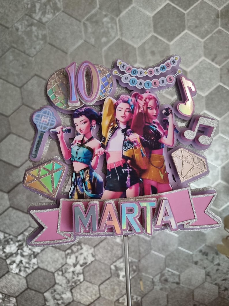 Puede incluir: Un adorno para tarta con tres figuras femeninas estilizadas sosteniendo micr&oacute;fonos. El adorno incluye el n&uacute;mero "10", el nombre "MARTA" y el texto "KPOP DEMON HUNTERS". El dise&ntilde;o incorpora acentos rosas, morados e iridiscentes.
