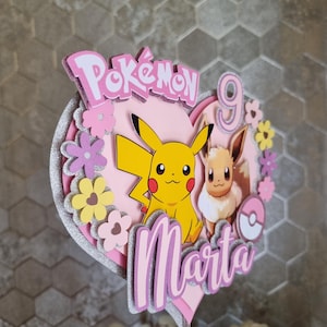 May include: Pink and yellow Pokemon cake topper featuring Pikachu and Eevee. The topper is shaped like a heart and has the words "Pokemon" and "Marta" on it. The number 9 is also on the topper.