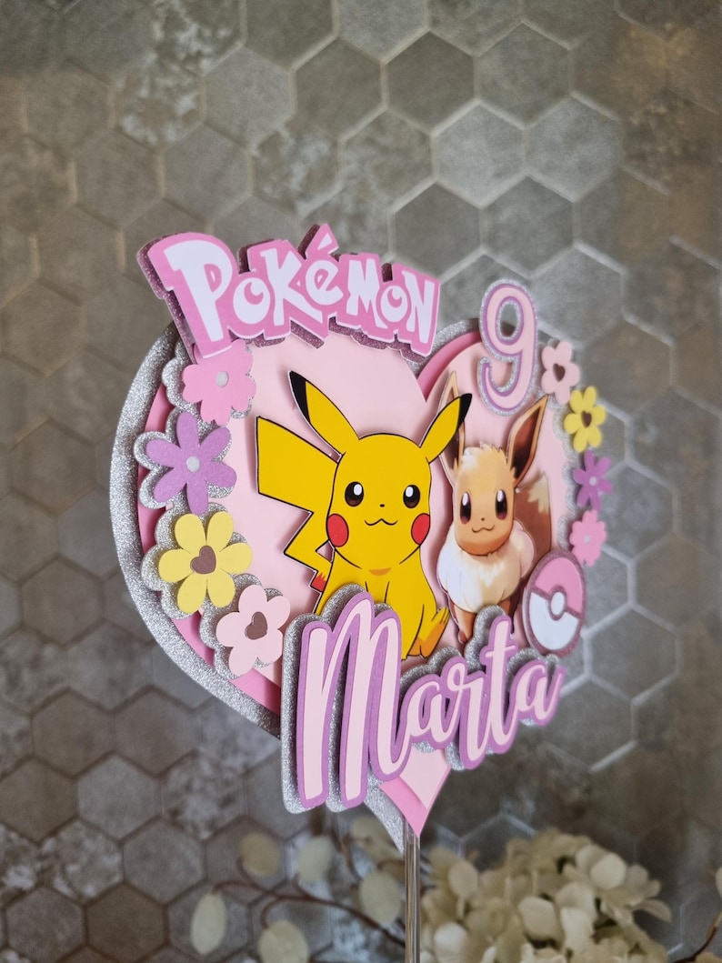 May include: Pink and yellow Pokemon cake topper featuring Pikachu and Eevee. The topper is shaped like a heart and has the words "Pokemon" and "Marta" on it. The number 9 is also on the topper.