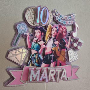 Puede incluir: Un adorno para pastel en capas con tres figuras estilizadas sosteniendo micr&oacute;fonos. El adorno incluye el n&uacute;mero "10", el nombre "MARTA" y notas musicales. El dise&ntilde;o incorpora detalles en rosa, morado y plata brillante, con las palabras "KPOP IDOL HUNTERS".