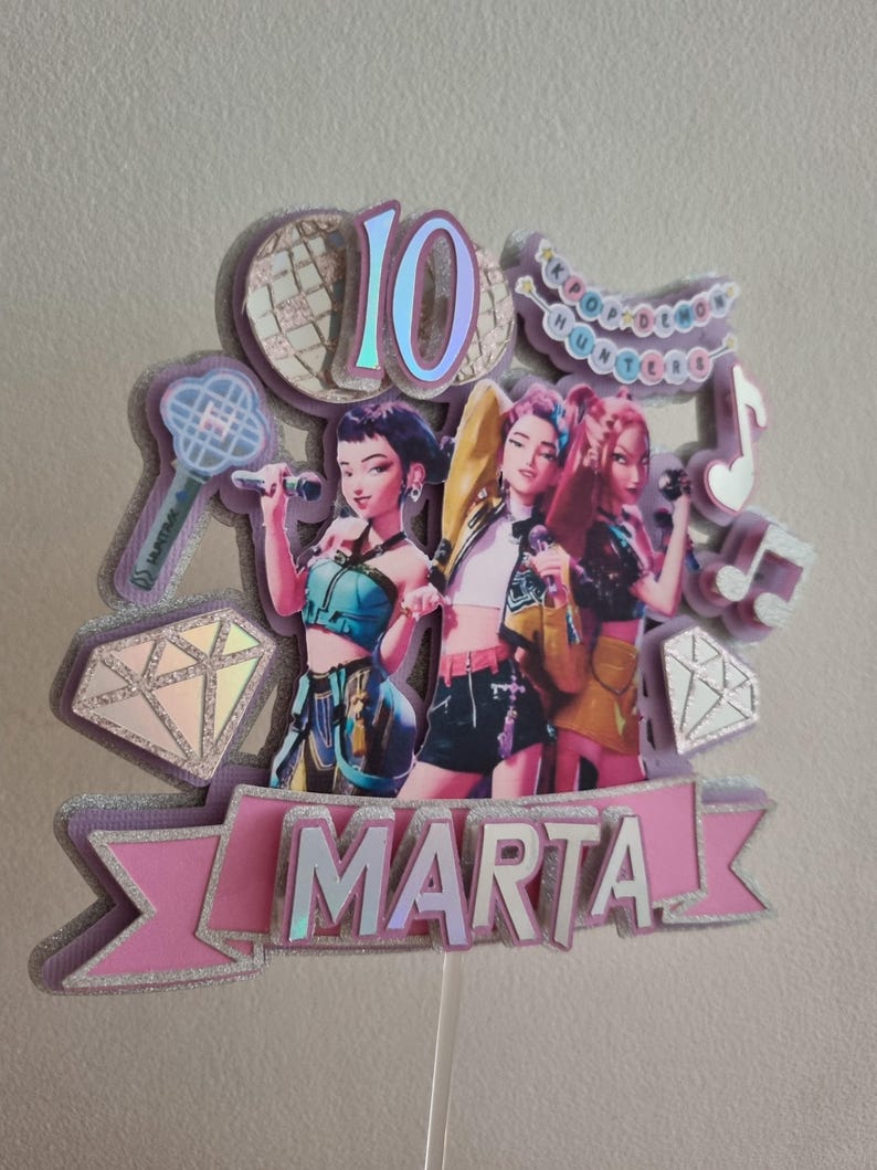 Puede incluir: Un adorno para pastel en capas con tres figuras estilizadas sosteniendo micr&oacute;fonos. El adorno incluye el n&uacute;mero "10", el nombre "MARTA" y notas musicales. El dise&ntilde;o incorpora detalles en rosa, morado y plata brillante, con las palabras "KPOP IDOL HUNTERS".