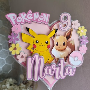 May include: A pink heart-shaped cake topper with a pink and white Pok&eacute; Ball, Pikachu and Eevee characters, the word "Pok&eacute;mon" and the name "Marta" in pink glitter letters, and the number "9" in pink glitter.