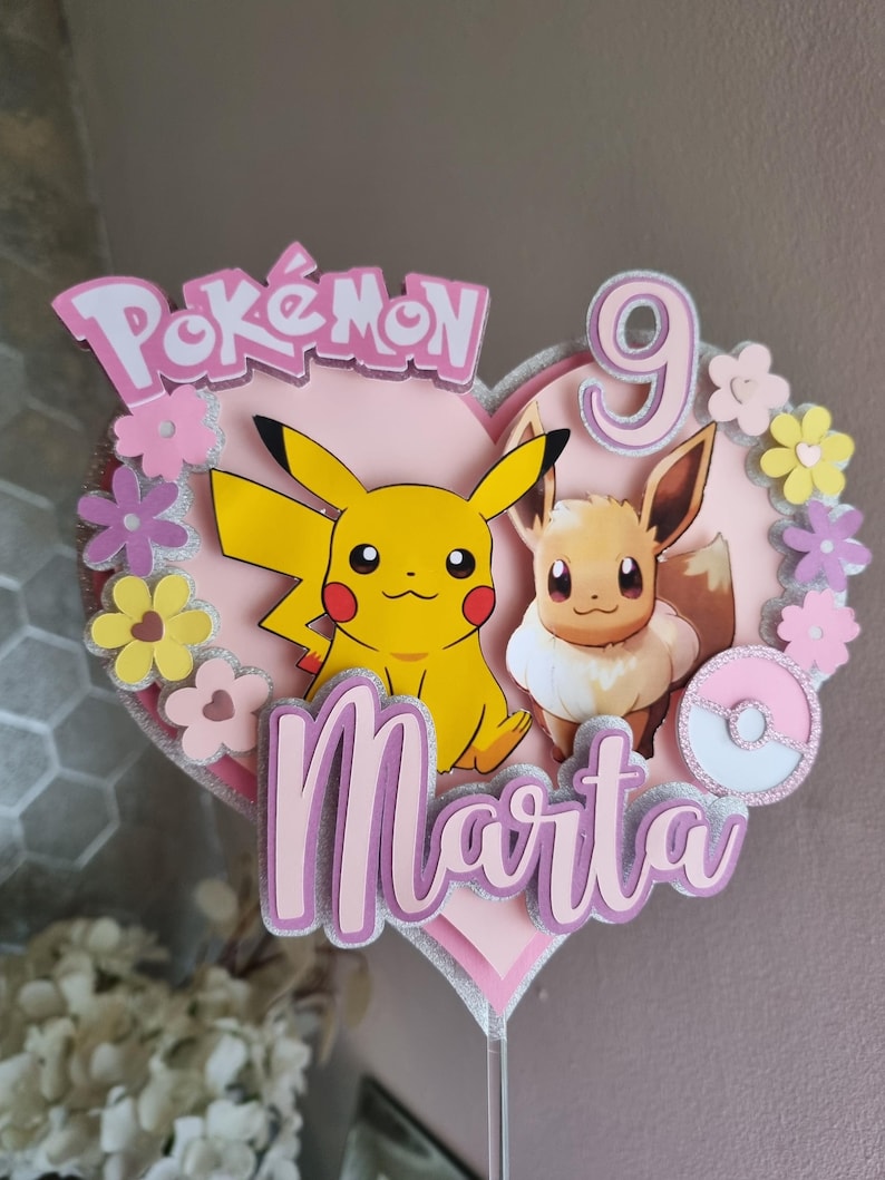 May include: A pink heart-shaped cake topper with a pink and white Pok&eacute; Ball, Pikachu and Eevee characters, the word "Pok&eacute;mon" and the name "Marta" in pink glitter letters, and the number "9" in pink glitter.
