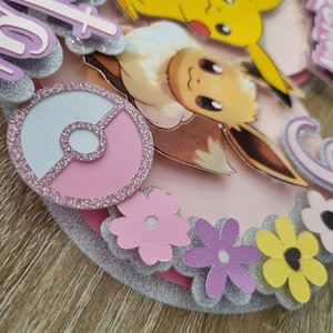 May include: A pink and silver cake topper featuring a pink Pok&eacute; Ball, a brown Eevee, a yellow Pikachu, and pink flowers. The cake topper reads "Happy 6th Birthday".