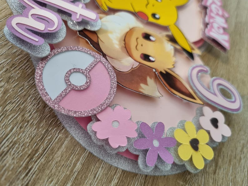 May include: A pink and silver cake topper featuring a pink Pok&eacute; Ball, a brown Eevee, a yellow Pikachu, and pink flowers. The cake topper reads "Happy 6th Birthday".