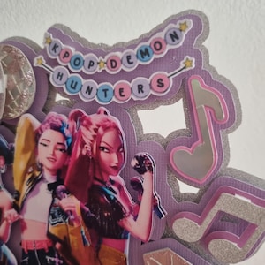 Puede incluir: Decoraci&oacute;n K-Pop en capas morada y plateada con el texto "KPOP DEMON HUNTERS". El dise&ntilde;o presenta ilustraciones de figuras animadas, notas musicales y detalles de estrellas.