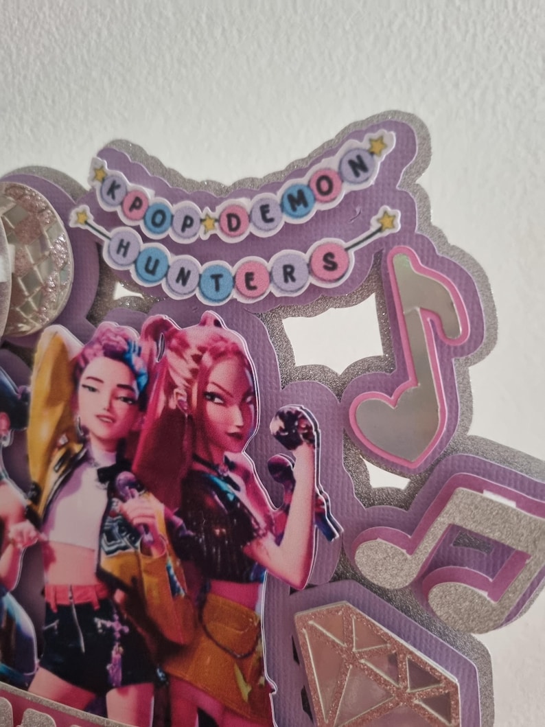 Puede incluir: Decoraci&oacute;n K-Pop en capas morada y plateada con el texto "KPOP DEMON HUNTERS". El dise&ntilde;o presenta ilustraciones de figuras animadas, notas musicales y detalles de estrellas.