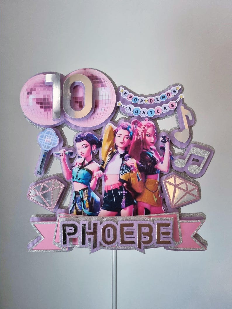Puede incluir: Adorno para tarta con tres figuras de dibujos animados sosteniendo micr&oacute;fonos. El adorno incluye el n&uacute;mero 10, el nombre "PHOEBE" y el texto "KPOP DEMON HUNTERS". El dise&ntilde;o incorpora detalles de purpurina rosa, plateada y morada.