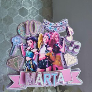 Puede incluir: Un adorno para tarta de varias capas con tres figuras estilizadas con micr&oacute;fonos. El adorno incluye el n&uacute;mero "10", notas musicales y el texto "KPOP DEMON HUNTERS". El nombre "MARTA" se muestra de forma destacada en una pancarta.