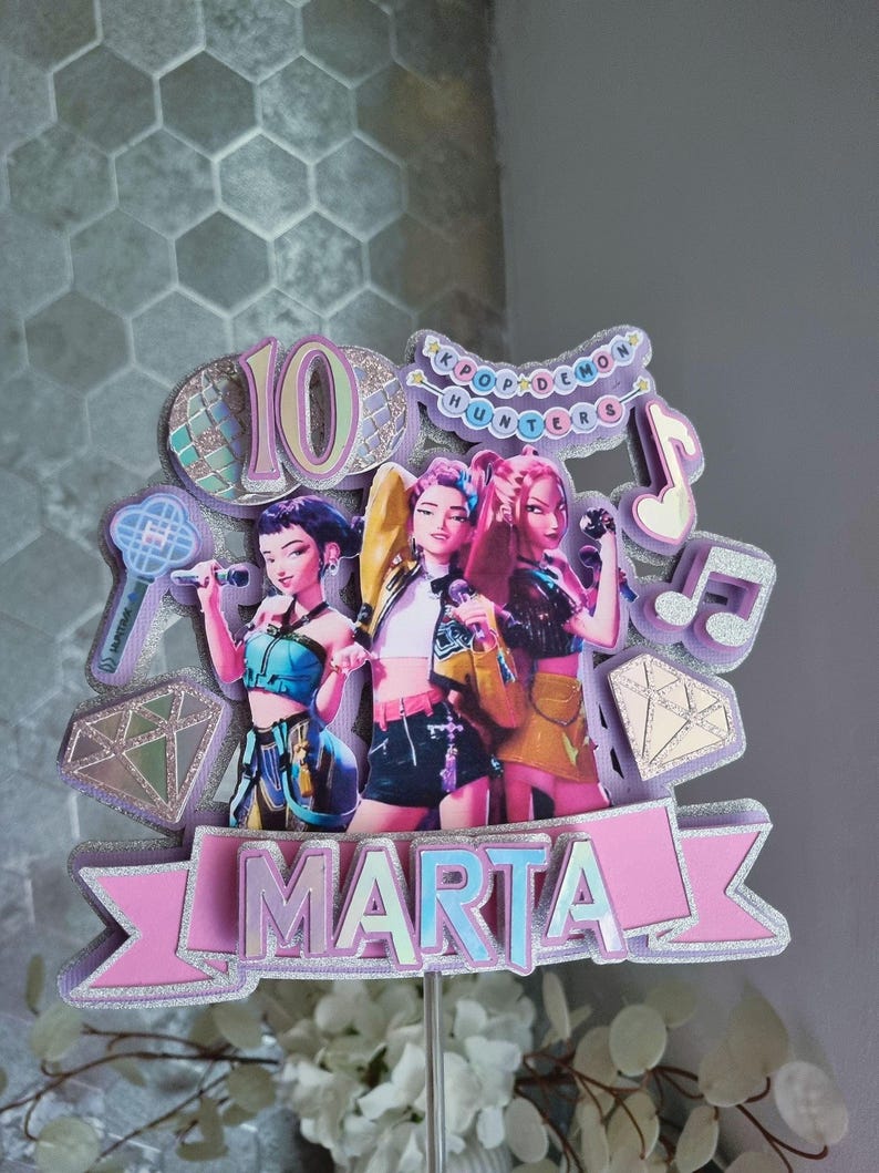 Puede incluir: Un adorno para tarta de varias capas con tres figuras estilizadas con micr&oacute;fonos. El adorno incluye el n&uacute;mero "10", notas musicales y el texto "KPOP DEMON HUNTERS". El nombre "MARTA" se muestra de forma destacada en una pancarta.