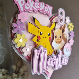 May include: A pink heart-shaped cake topper featuring Pikachu and Eevee characters, the words "Pok&eacute;mon" and "Marta", and the number 9. The topper is decorated with pink, yellow, and white flowers.