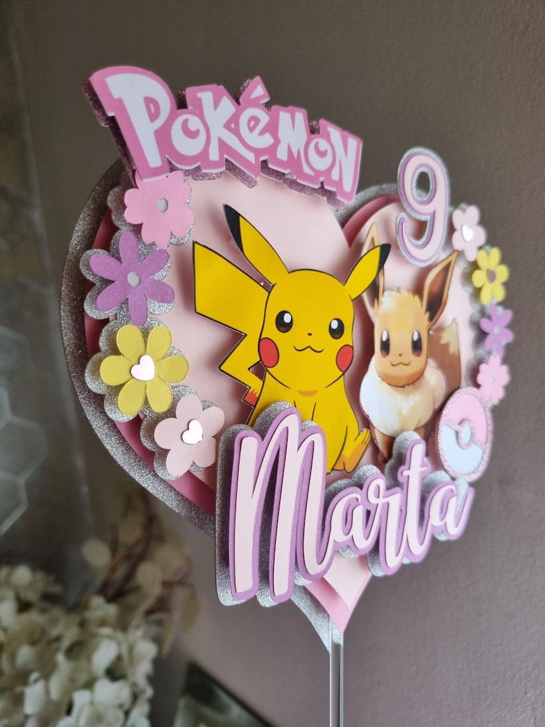 May include: A pink heart-shaped cake topper featuring Pikachu and Eevee characters, the words "Pok&eacute;mon" and "Marta", and the number 9. The topper is decorated with pink, yellow, and white flowers.