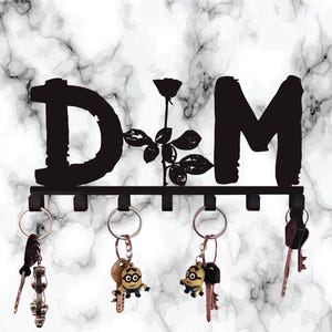 Puede incluir: Llavero de metal negro con las letras "D" y "M" conectadas por una rosa. El llavero tiene seis ganchos con llaves y llaveros colgando. El fondo es un patrón de mármol blanco y gris.