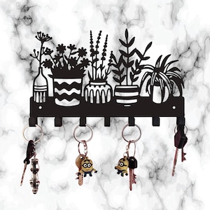 May include: Black metal key holder featuring a decorative silhouette of potted plants. The key rack has six hooks and is mounted on a white and gray marble-patterned background. Several keys with keychains are hanging from the hooks.