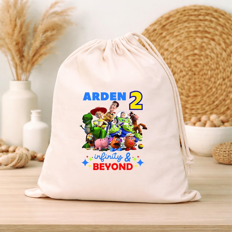 K&ouml;nnte beinhalten: Ein beiger Kordelzugbeutel mit einer farbenfrohen Grafik von Figuren aus dem Animationsfilm "Toy Story". Der Beutel tr&auml;gt den Namen "ARDEN" und die Zahl "2" in Blau und Gelb sowie den Schriftzug "infinity & BEYOND."